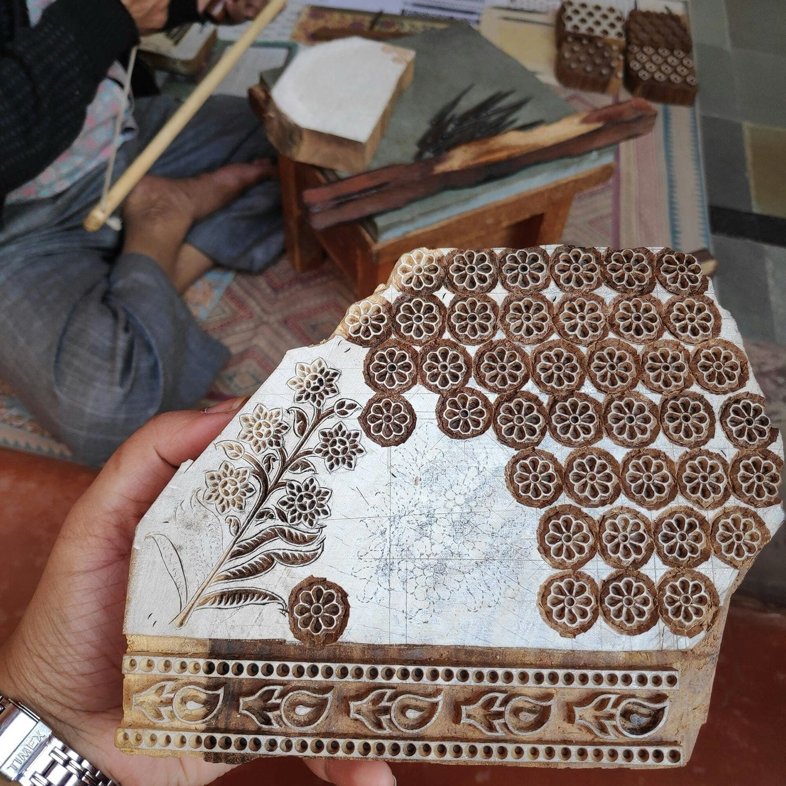 A hand holding a carved wooden textile-printing block, its face cut with rows of flower motifs and a patterned border.
