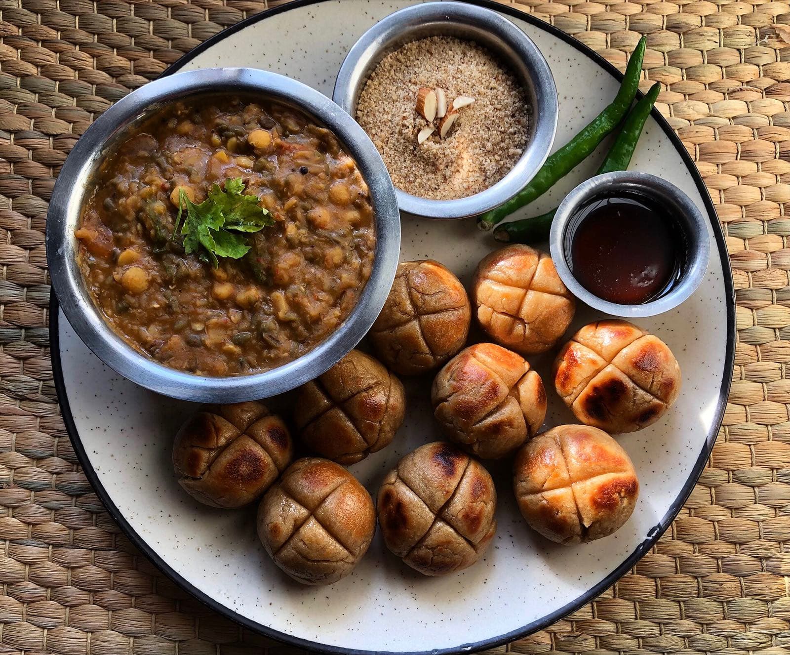 A plate of dal baati churma: a bowl of lentil dal, round baked wheat baati, a bowl of crumbled churma and green chillies.